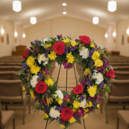 Standing wreath of red roses, yellow mums, and white flowers on an easel in a chapel.
