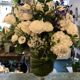 Tall arrangement of white roses and hydrangeas with purple delphinium in a clear glass pedestal vase