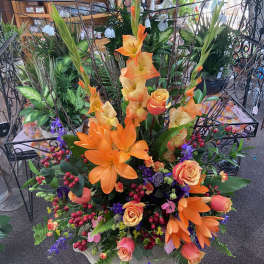 Tall arrangement of orange lilies and gladiolus with peach roses and purple flowers in a mixed display