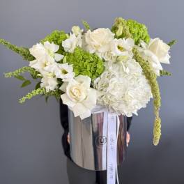 White and green flower arrangement with roses and hydrangeas in a tall silver hatbox-style container