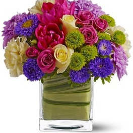 Mixed bouquet of pink, purple, and cream flowers in a clear square vase