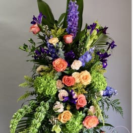 Tall standing arrangement of orange roses, purple delphinium, irises, and green hydrangeas with bold tropical foliage.