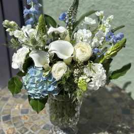 Arrangement of white calla lilies, roses, and blue hydrangea in a tall clear glass vase