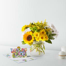 Yellow and white flowers in a glass vase beside a "Thinking of You" card