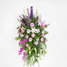 Tall standing spray of lavender roses, pink carnations, white lilies, and purple blooms on an easel.