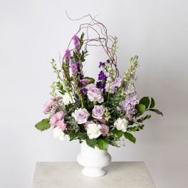 Tall arrangement of lavender roses and purple blooms in a white pedestal vase