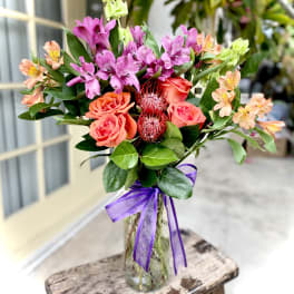 Mixed bouquet of pink and orange flowers in a glass vase with a purple ribbon
