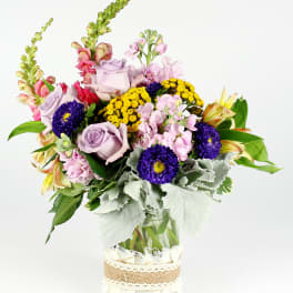 Mixed bouquet of lavender roses, pink blooms, and yellow flowers in a glass vase