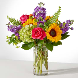 Mixed bouquet of sunflowers, pink roses, and purple blooms in a glass vase