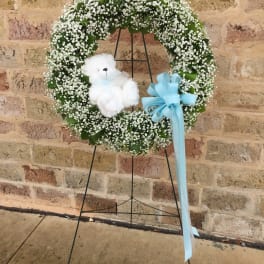 White baby’s breath wreath with white teddy bear and blue bow on a stand against a brick wall