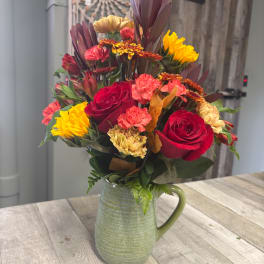 Mixed bouquet of red roses, carnations, and sunflowers in a green ceramic pitcher