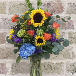 Mixed bouquet with sunflowers, orange roses, and blue hydrangea in a glass vase