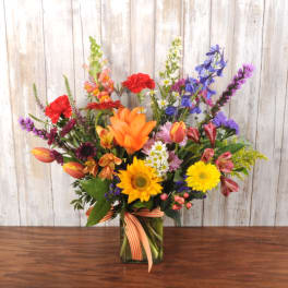 Colorful mixed bouquet with sunflowers, lilies, tulips, and wildflowers in a glass cube vase with ribbon