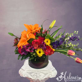 Colorful bouquet with orange lilies, red roses, yellow daisies, and purple flowers in a dark vase