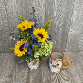 Yellow sunflowers and blue blooms arranged in a ceramic owl vase on a wood surface