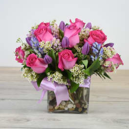Compact arrangement of pink roses and purple flowers in a square glass vase with stones and a satin ribbon.