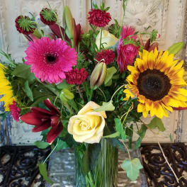 Mixed bouquet with sunflowers, roses, lilies, and pink gerbera daisies in a glass vase