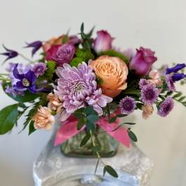 Low arrangement of purple mums, peach roses, and mixed blooms in a glass vase with a pink ribbon.
