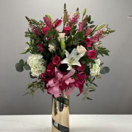 Tall bouquet of pink roses and white lilies in a gold vase with a pink ribbon