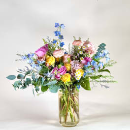 Tall mixed bouquet of pink, yellow, white, and blue flowers arranged in a clear glass vase