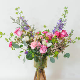 Tall mixed arrangement of pink roses, carnations, and purple flowers in a ribbed glass vase