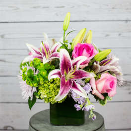 Compact arrangement of pink lilies and roses with green accents in a square glass vase.