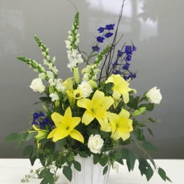 Yellow lilies and white roses arranged in a white basket with blue flowers.
