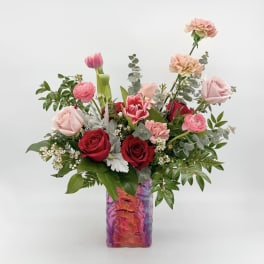 Mixed pink and red roses, tulips, ranunculus, and carnations in a colorful glass vase