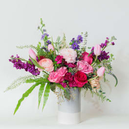 Asymmetrical pink and purple flower arrangement with tulips and roses in a tall white and gray cylinder vase