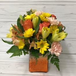 Mixed bouquet of yellow, peach, and coral flowers in a square glass vase