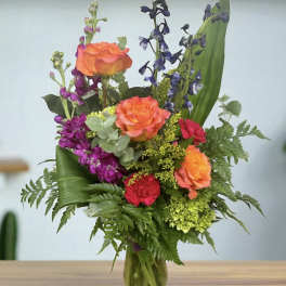 Mixed bouquet of orange roses, purple flowers, and greenery in a glass vase