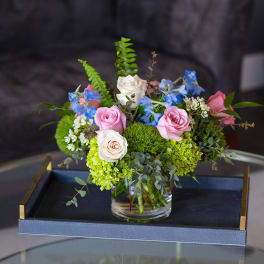 Low arrangement of pink and ivory roses with blue flowers in a clear glass vase