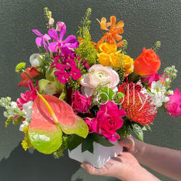Bright mixed arrangement of pink, yellow, and orange flowers in a white cube vase.