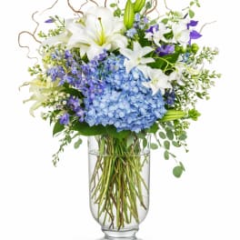 Tall arrangement of white lilies and blue hydrangeas with purple accents in a clear glass vase