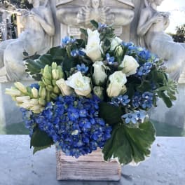Compact blue and white flower arrangement with hydrangeas and roses in a rustic wooden cube vase.