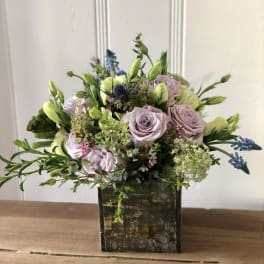 Lavender roses and white buds arranged with blue accents in a rustic wooden cube vase