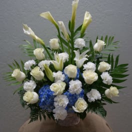 White calla lilies, roses, and carnations arranged with blue hydrangeas in a basket