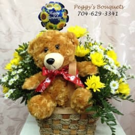 Teddy bear in a basket surrounded by yellow and white flowers with a birthday balloon