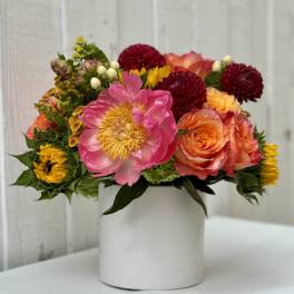 Bright arrangement of pink peony, orange roses, and yellow blooms in a white cylinder vase