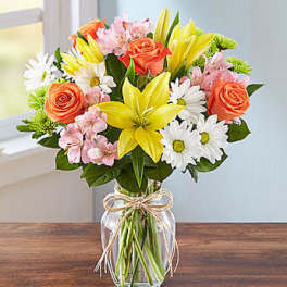 Bouquet of yellow lilies, orange roses, pink flowers, and white daisies in a glass vase
