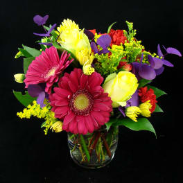 Mixed bouquet of pink gerbera daisies, yellow roses, and purple accents in a glass vase