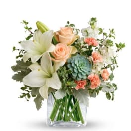 Bouquet of white lilies, peach roses, and blue-green blooms in a clear vase