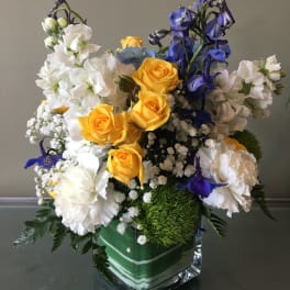 Compact arrangement of yellow roses, white blooms, and blue flowers in a square glass vase