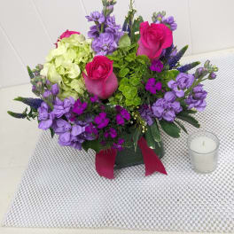 Compact arrangement of hot pink roses, green hydrangea, and purple blooms in a glass vase with ribbon