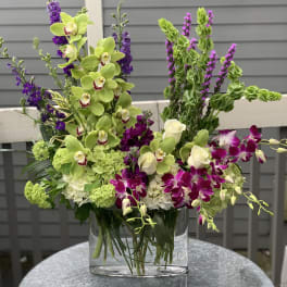 Tall floral arrangement with green orchids, purple blooms, and white roses in a glass vase