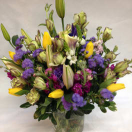Mixed bouquet of yellow tulips, purple blooms, and lilies in a glass vase