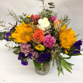 Mixed bouquet with sunflowers, roses, and purple filler in a glass vase