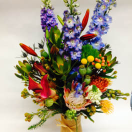Tall mixed bouquet with blue-purple flowers and red lilies in a burlap-wrapped vase