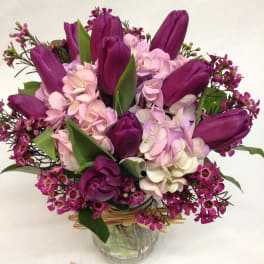 Purple tulips with pink hydrangeas and small blooms in a clear glass jar vase tied with raffia.