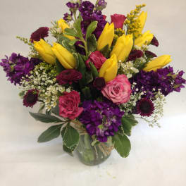 Bouquet of yellow tulips, pink roses, and purple flowers in a glass vase